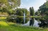Serene park landscape featuring a reflective pond with a central fountain surrounded by lush greenery and tall trees.