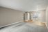 Bright open living area with neutral tones, carpeted floor, and a glimpse of a modern kitchen with hardwood flooring.