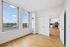 Bright apartment interior with large windows offering cityscape views, leading to a modern kitchen featuring sleek white cabinetry and stainless steel appliances.