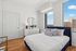 Bright and modern bedroom featuring a cozy bed, elegant dresser, and a stunning cityscape view through generous windows.