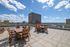 Spacious rooftop patio featuring modern wooden furniture, offering stunning cityscape views under a bright blue sky with fluffy clouds.