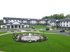 Charming apartment complex with lush green lawns, a decorative fountain centerpiece, and ample parking space, offering a welcoming community vibe.