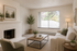 Elegant living room with a cozy white couch, modern coffee table, and a stylish fireplace, bathed in natural light from large windows, featuring tasteful decor and greenery.