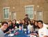 Group of friends enjoying a vibrant outdoor meal, seated around a blue table against a modern brick building backdrop.