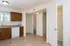 Empty kitchen with beige tile flooring, white walls, and brown cabinets. Open door reveals hallway with soft lighting, creating a clean, neutral atmosphere.