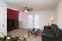 A cozy living room with a black leather sofa, glass coffee table, and TV cabinet against a red accent wall. Ceiling fan above and soft lighting create a warm atmosphere.