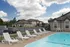 A row of white lounge chairs lines the fenced-in pool area at Chesterfield Village Apartments. A beige clubhouse with large windows is adjacent to the pool, with apartment buildings and a parking lot visible in the background under a partly cloudy sky.