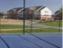 Blue tennis court with white lines in the foreground, enclosed by a black chain-link fence. In the background, there are residential buildings under a clear blue sky, conveying a calm and suburban atmosphere.