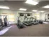 A well-lit gym with two treadmills, an elliptical machine, and two wall-mounted TVs. Wooden blinds cover the windows, creating a clean, inviting atmosphere.