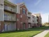 Three-story apartment building with red brick and beige siding, white balconies, and a well-kept lawn under a clear blue sky. Peaceful suburban setting.