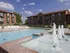 Clear daytime view of a swimming pool and spa area in an apartment complex. The pool is surrounded by lounge chairs, and a fountain feature with multiple water jets flows in the foreground. Brick apartment buildings with balconies and green awnings encircle the pool area, with a blue sky and scattered white clouds overhead, creating a relaxing outdoor environment.
