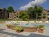 Daytime view of an apartment complex pool area featuring a brick fountain with multiple water jets in the foreground, surrounded by potted plants with lush greenery. White lounge chairs line the poolside, and brick apartment buildings with green awnings frame the background. The sky is clear with a few clouds, giving a calm and sunny atmosphere.
