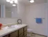 Bright bathroom with a large mirror above a wooden vanity, featuring a single sink and a beige countertop. Decor includes a clock, a small plant, and a blue towel hanging on the wall. The shower area is partially visible in the background, and overhead lighting provides a warm glow.