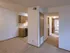 Hallway view of an apartment with carpeted flooring, leading from the living area toward the bedrooms and bathroom. The kitchen with wooden cabinets is visible on the left, giving a sense of open layout and accessibility.