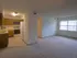 Spacious living area with an open layout that connects to the kitchen. The room is carpeted, and a large window on the right lets in natural light. The kitchen features wooden cabinetry and standard appliances.