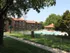 Daytime view of an apartment complex with a pool area enclosed by a black metal fence. The scene features a grassy area with a sidewalk and mature trees providing shade in the foreground. The brick buildings with green awnings surround the pool, creating a serene and inviting atmosphere.