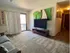Living room with carpeted flooring, featuring a large TV on a wooden stand, artwork on the walls, and a sliding glass door leading to a private balcony. The space is furnished with a comfortable gray couch and decorated with personalized touches.