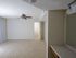 Spacious empty room with white walls and beige carpet. Ceiling fan and light are visible, with an open door leading to another room. Bright and clean ambiance.