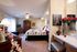Cozy bedroom with a floral duvet on a bed, wooden armoire, and a ceiling fan; a desk with a Paris-themed artwork and vase of flowers adds elegance.