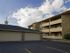 Two beige buildings under a partly cloudy sky, featuring a three-story apartment complex and a one-story garage with stone accents. Calm, suburban setting.