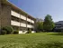A three-story brick apartment building with balconies is surrounded by a well-kept lawn and lush trees under a clear blue sky, conveying a peaceful suburban vibe.