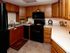 Warm, inviting kitchen with wooden cabinets, white countertops, black appliances, and a tiled floor. A clock and plant add decorative touches.