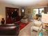 Cozy living room with beige couches, large wooden entertainment center, grandfather clock, red rug, and glass doors leading to a sunny patio with greenery.