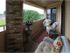 A cozy veranda with wicker chairs and floral cushions on a woven rug. A large vase and potted plants sit near a brick wall, creating a tranquil ambiance.