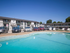 Large outdoor swimming pool surrounded by a two-story apartment building with white siding and parked vehicles, set beneath a clear blue sky.