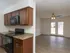 A modern apartment kitchen with dark wood cabinetry, black appliances, and a beige speckled countertop. The kitchen opens to a spacious living area with light brown wood-style flooring. A ceiling fan with lights is centered in the living space, and French doors with white trim lead to an outdoor patio.