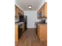 A compact galley-style kitchen with warm wood cabinetry and black appliances, including a stove, microwave, refrigerator, and dishwasher. The brown speckled countertops contrast with the white walls. A single light fixture illuminates the space, and the hardwood-style flooring extends throughout.