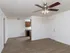 A large living room with wood-style flooring and a ceiling fan with a central light fixture. The kitchen is visible in the background, featuring dark wood cabinetry and black appliances. A white entry door is located on the right side of the room.