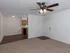 A bright, open-concept living area with beige carpeting and a white ceiling fan with wooden blades. The kitchen, visible in the background, features warm wood cabinets and dark appliances. A white entry door is adjacent to the kitchen space.