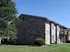 A two-story brick building with large windows is surrounded by a well-kept lawn and a tree on a sunny day, conveying a sense of tranquility.