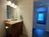 Bathroom with a wooden vanity, white countertop, and sink; lit mirror above. Blue towel hung nearby. Open door reveals bedroom with natural light.