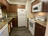A narrow L-shaped kitchen with wood cabinets, white appliances, and dark laminate flooring. It has a cozy feel with a dishwasher and white towels hanging.