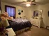 Cozy bedroom with a large bed covered in a purple floral comforter and plush toys. A ceiling fan spins above, and a white dresser with a mirror stands nearby.