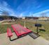Accessible concrete picnic area with a red metal table and bench, adjacent to a black grill and surrounded by grassy lawn within a fenced community area.