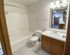Bathroom with white acrylic bathtub and shower combo, standard toilet, and a wood vanity with white countertop and single sink. A large mirror with overhead lighting is mounted above the sink, and the room has light-colored tile flooring. A towel bar is installed on the wall beside the toilet.