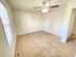 Spacious, empty room with cream-colored walls, beige carpet, and a ceiling fan with lights. A window on the left side allows natural light in.
