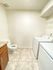 A small laundry room with a washing machine and dryer on the right. To the left, a toilet and a wooden cabinet with marble top. The space is clean and simple.