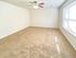 An empty room with light beige carpet, white walls, and a ceiling fan with red blades. Sunlight streams through a large window, creating a bright, airy ambiance.