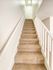A carpeted staircase with beige tones ascends between two white walls, illuminated by soft natural light. Wooden handrails run alongside, creating a clean, inviting atmosphere.