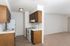 Compact kitchen with wooden cabinets, white appliances, and tiled flooring, leading into a spacious, empty living area.