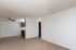 Spacious living area with beige carpet, white walls, and open layout leading towards a kitchen area in the background.