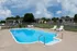 Fenced outdoor swimming pool with clear blue water, white lounge chairs, and a clean concrete deck, surrounded by grassy areas and red brick apartment buildings in the background.