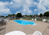 Wide view of pool area showing full layout with lounge chairs, plastic seating, safety railing, and chain-link fence enclosing the space with multiple apartment buildings visible beyond.
