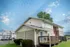 Suburban house with beige siding and a small wooden deck, set against a clear blue sky with scattered clouds. Lush green bushes and a tree are in the background.