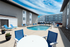 Outdoor pool area with lounge chairs and a round table with blue chairs under a bright sky. Surrounded by modern buildings, conveying a relaxing vibe.