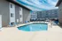 Modern apartment complex with a clear blue swimming pool, surrounded by lounge chairs. Buildings with large windows under a partly cloudy sky